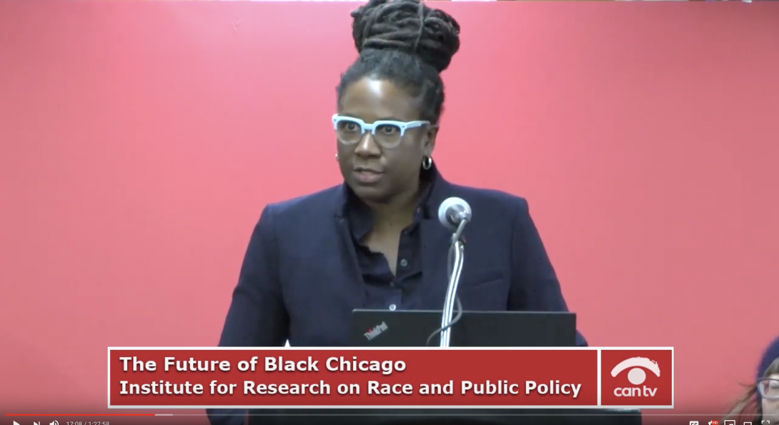 a black woman with blue glasses and her hair pulled up stands at a podium speaking