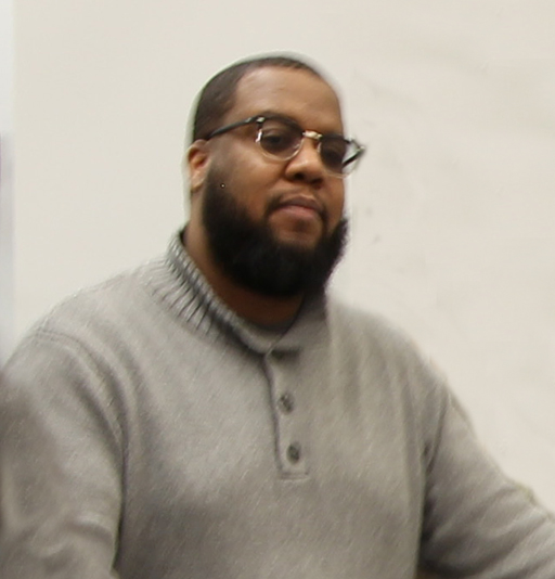 Man in grey sweater, glasses, and a beard looks at the camera
