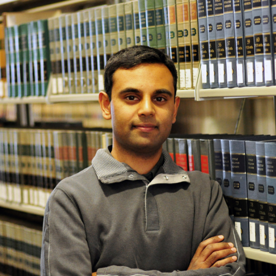man in grey shirt and arms crossed looks at the camera in front of a row of library books