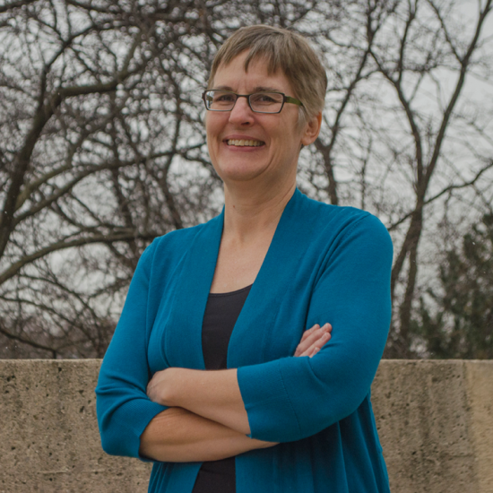 woman in blue sweater, short hair and glasses standing outside smiles for the camera