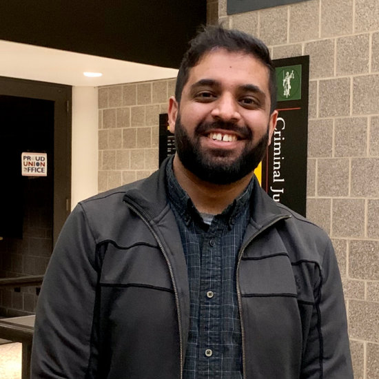 A man with a beard and short hair wearing a brown jacket stands in a hallway at UIC and smiles before the camera