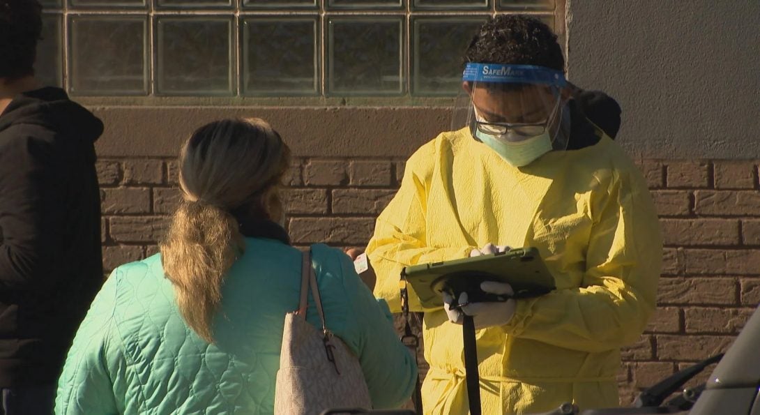 Photograph of health worker in full yellow protective suit, face mask, and face shield writing information into a tablet from a woman in a green jacket who stands in front of her