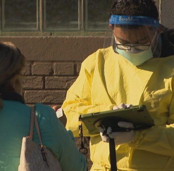 Photograph of health worker in full yellow protective suit, face mask, and face shield writing information into a tablet from a woman in a green jacket who stands in front of her 