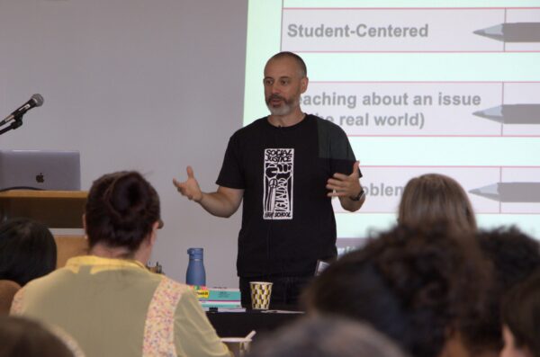 A person in a black t-shirt with a white graphic on the front stands at the front of an audience. He is gesturing with his hands.